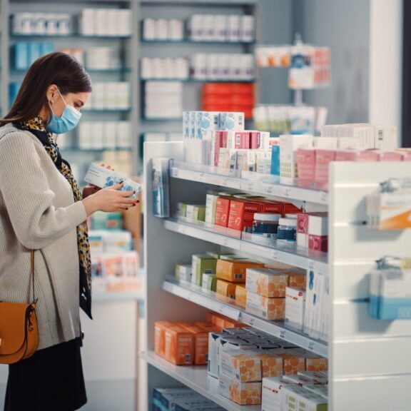 Pharmacy Drugstore: Portrait of Beautiful Young Woman Wears Protective Face Mask, Searches to Buy Best Medicine, Drugs, Vitamins. Shelves full of Health Care, Welness, Cosmetics, Beauty Products